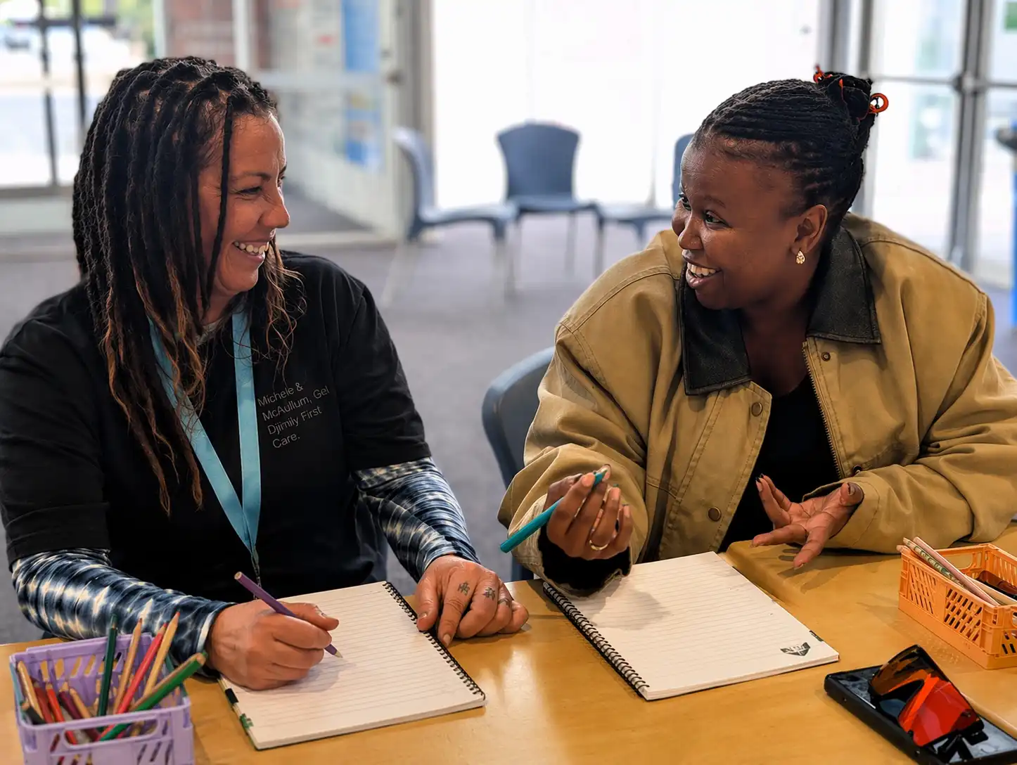 Two women sitting at a table during a Mandurah Disability Network meeting, smiling and talking while taking notes, with pens and notebooks on the table